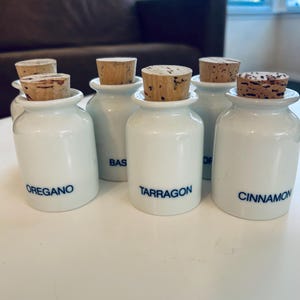 Può includere: Una collezione di vasetti di spezie in ceramica bianca con tappi in sughero. Ogni vasetto è etichettato con un nome di spezia diverso in testo blu: Origano, Dragoncello e Cannella. I vasetti sono disposti su una superficie bianca.