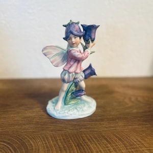 May include: A porcelain figurine of a fairy holding two large bluebell flowers. The fairy wears a pink top, blue shorts, and a matching hat. The fairy has delicate wings and stands on a small, round base with blue and green accents.
