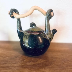 Puede incluir: Una tetera de cerámica hecha a mano con un cuerpo oscuro y moteado y un asa curva de color claro. La tetera presenta una pequeña tapa y un diseño artístico único. La paleta de colores incluye tonos negros, marrones y beige.