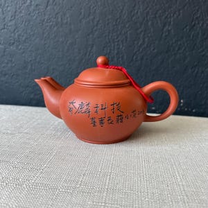 May include: A small, reddish-brown ceramic teapot with a rounded body, a curved spout, and a loop handle. The lid is topped with a small knob and a red string. The teapot has black Chinese characters on its side.