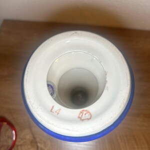 May include: A white and blue ceramic vase with a wide rim. The rim has a blue band and the text "L4" is visible. The vase has a circular opening and a dark interior. A red object is visible in the lower left corner.
