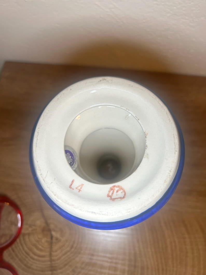May include: A white and blue ceramic vase with a wide rim. The rim has a blue band and the text "L4" is visible. The vase has a circular opening and a dark interior. A red object is visible in the lower left corner.