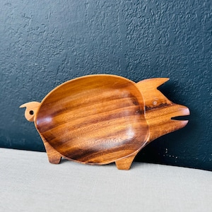 Puede incluir: Un plato de servir de madera con forma de cerdo. El plato es de madera marrón oscura y tiene una veta de madera natural. El cerdo tiene una cola pequeña y un hocico.