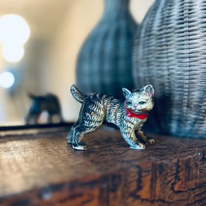 May include: A ceramic cat figurine with a red collar stands on a wooden surface. The cat has a detailed, patterned coat in shades of gray, black, and tan. Two decorative vases are in the background.