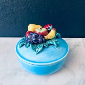 May include: A blue ceramic trinket box with a lid featuring a 3D design of fruit, including a pear, apple, grapes, banana, and strawberries.