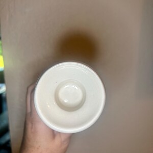 May include: A white ceramic candle holder with a circular design. The candle holder has a small, raised center and a wide, flat rim. The holder is held by a hand.