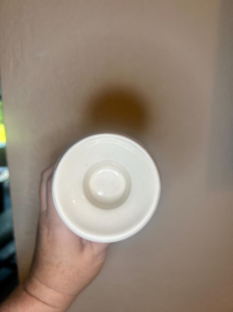 May include: A white ceramic candle holder with a circular design. The candle holder has a small, raised center and a wide, flat rim. The holder is held by a hand.