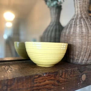 Puede incluir: Un cuenco de cerámica amarillo claro con líneas horizontales, reflejado en un espejo. El cuenco está sobre una superficie de madera oscura, con un jarrón tejido y vegetación en el fondo. El cuenco es de color amarillo claro.