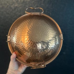 May include: A round, hammered copper bowl with a metallic finish. The bowl features a prominent, textured surface with small, concave indentations throughout. It includes a small, arched metal handle attached to the top rim and metal hinges on the sides.