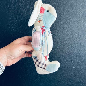 May include: A stuffed rabbit toy constructed from various fabric patches in shades of blue, red, and white. The plush features a small black bead eye, long floppy ears, and a thin blue ribbon tied around its neck. The body displays a mix of solid, checkered, and floral patterns.
