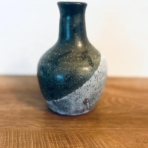 Puede incluir: Un pequeño jarrón de cerámica hecho a mano con un esmalte moteado verde oscuro y blanco. El jarrón tiene un cuerpo redondeado y un cuello estrecho, y mide aproximadamente 15 cm de alto. El jarrón se exhibe sobre una superficie de madera.