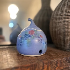 Op de afbeelding: Een lichtblauwe keramische etherische olie diffuser met een bloemenontwerp in rood, blauw en groen. De diffuser heeft een kleine opening en een gebogen bovenkant. Hij staat op een houten oppervlak.