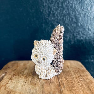 Puede incluir: Una figura de cerámica de ardilla con un cuerpo texturizado blanquecino y una cola marrón texturizada. La ardilla tiene ojos pequeños, redondos y amarillos y está en posición vertical. La figura se exhibe sobre una superficie de madera sobre un fondo azul oscuro.