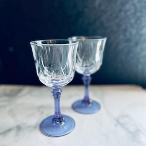 May include: Two clear crystal wine glasses with a decorative pattern on the bowl and a purple stem and base. The glasses are set against a dark blue background on a white marble surface.