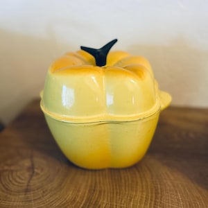 May include: A yellow ceramic pumpkin-shaped casserole dish with a black stem. The dish has a glossy finish and two small handles. The lid fits snugly on the base. The dish is on a wooden surface.