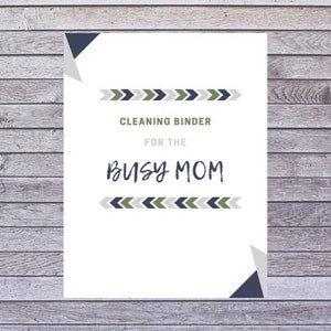 May include: A white paper with the text "CLEANING BINDER FOR THE BUSY MOM" in a mix of fonts. The text is framed by decorative arrow patterns in green, navy, and gray. The background is a wooden surface.