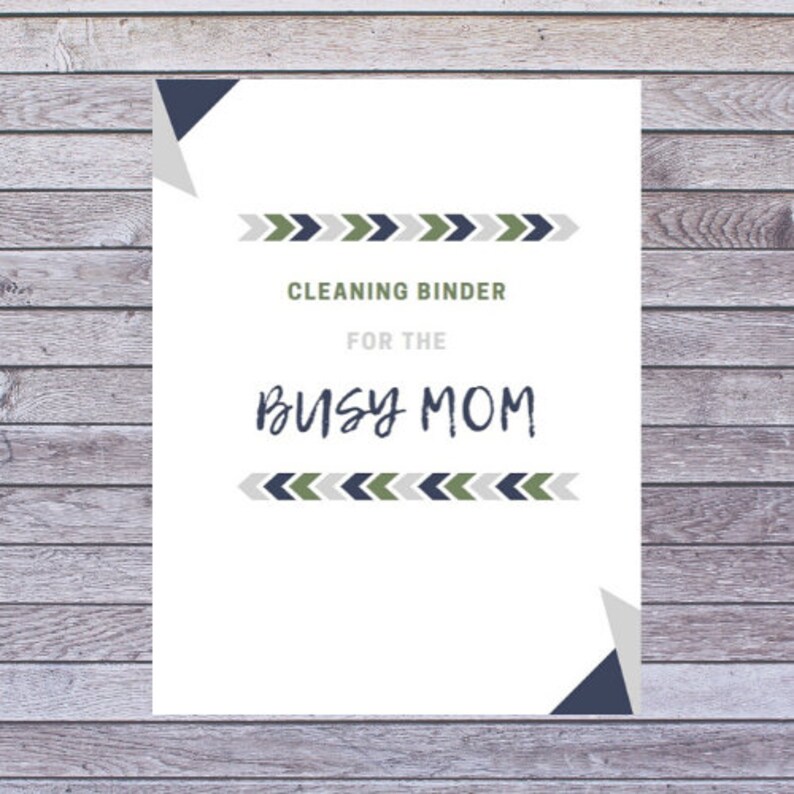 May include: A white paper with the text "CLEANING BINDER FOR THE BUSY MOM" in a mix of fonts. The text is framed by decorative arrow patterns in green, navy, and gray. The background is a wooden surface.