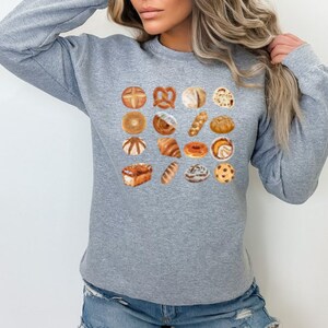 May include: A gray sweatshirt featuring a grid of various baked goods illustrations, including pretzels, bagels, croissants, and cookies. The design is printed on a soft, long-sleeved crew neck sweatshirt.
