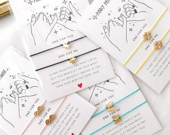 May include: Four sets of friendship bracelets with pink, yellow, black, and turquoise string. Each set has a gold charm with a star, infinity symbol, butterfly, or flower. The cards have the text "Pinky Promise..." and "One for you" and "One for me".