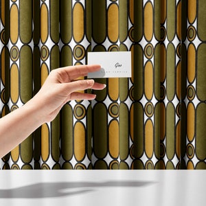 May include: A white business card with the text "Gina" and "www.ginafranklin.com" printed on it. The card is being held in front of a green and gold patterned curtain.