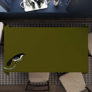 Könnte beinhalten: Eine grüne Tischdecke mit einer Illustration eines schwarz-weißen Vogels. Die Tischdecke liegt auf einem Tisch mit zwei Stühlen.