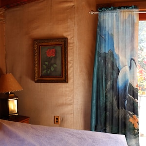 Può includere: Un interno di una camera da letto con una parete in legno, un letto con un copriletto bianco, una lampada su un tavolo in legno, un quadro incorniciato di una rosa rossa e una tenda blu e bianca con un disegno di un uccello.