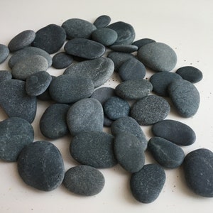 Puede incluir: Una colección de piedras de río lisas y grises de varios tamaños. Las piedras están dispuestas en una pila sobre una superficie blanca.