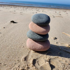 May include: A stack of four smooth stones, two gray and two pink, balanced on top of each other on a sandy beach.