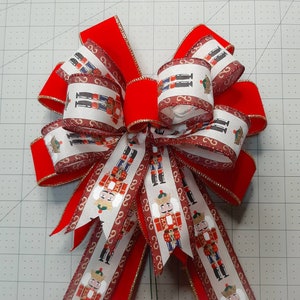 May include: A red and white bow with a gold trim. The bow is made of ribbon with a pattern of nutcracker soldiers. The bow is tied in a loop with the ends of the ribbon hanging down.