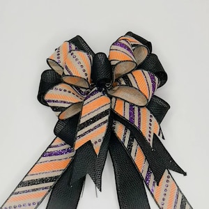 May include: A black and orange striped ribbon bow with purple glitter accents. The bow is tied with a black ribbon and is perfect for a Halloween wreath or door decoration.