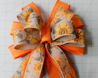 May include: A large, orange and brown bow made from burlap ribbon with a sunflower print. The bow is tied with a long, orange ribbon.