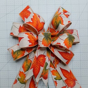 May include: A large, multi-layered bow made from beige burlap ribbon with a printed pattern of orange, green, and brown autumn leaves. The bow is tied with a loop at the top and has long tails.