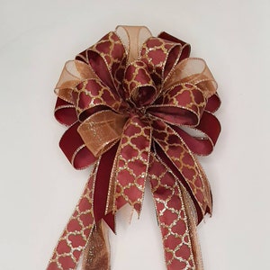 May include: A decorative bow featuring layers of burgundy and gold ribbons. The burgundy ribbons have a gold geometric pattern, while the gold ribbons are sheer. The bow is suitable for gift wrapping or holiday decorations.