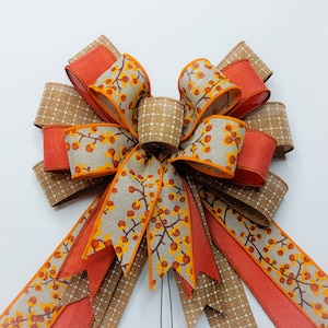 May include: A large, handmade bow with a brown and white gingham ribbon, orange ribbon, and a beige ribbon with a floral pattern. The bow is tied with a wire loop for hanging.