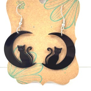 Peut inclure: Boucles d'oreilles chat noir avec un motif de croissant de lune. Les boucles d'oreilles sont en acrylique noir et présentent une silhouette de chat assis à l'intérieur d'une forme de lune incurvée. Les boucles d'oreilles sont suspendues à des crochets d'oreille argentés.