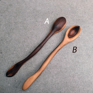 May include: Two wooden spoons labeled A and B. Spoon A is dark brown with a lighter brown handle. Spoon B is light tan with a dark brown circular grain pattern in the bowl. Both spoons feature long, slender, curved handles and smooth surfaces.