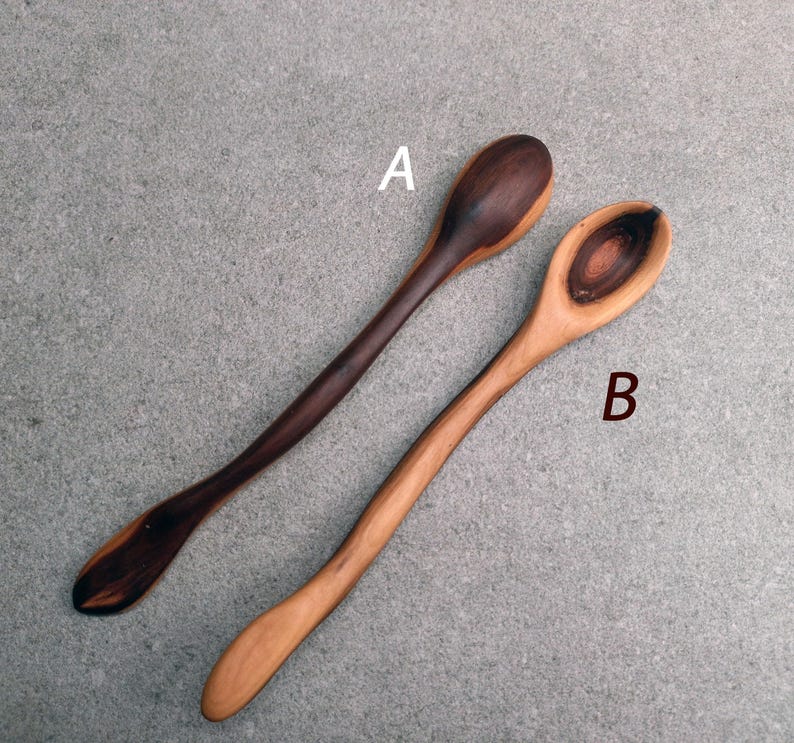 May include: Two wooden spoons labeled A and B. Spoon A is dark brown with a lighter brown handle. Spoon B is light tan with a dark brown circular grain pattern in the bowl. Both spoons feature long, slender, curved handles and smooth surfaces.