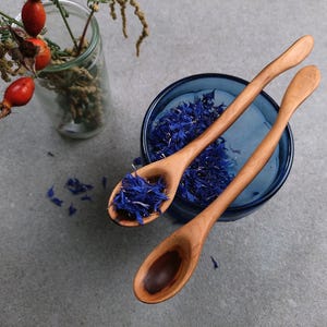 May include: Two light brown wooden spoons with curved handles. One spoon rests inside a dark blue glass bowl filled with dried blue cornflower petals, while the other spoon lies beside it. A clear glass vase with red rose hips is nearby.