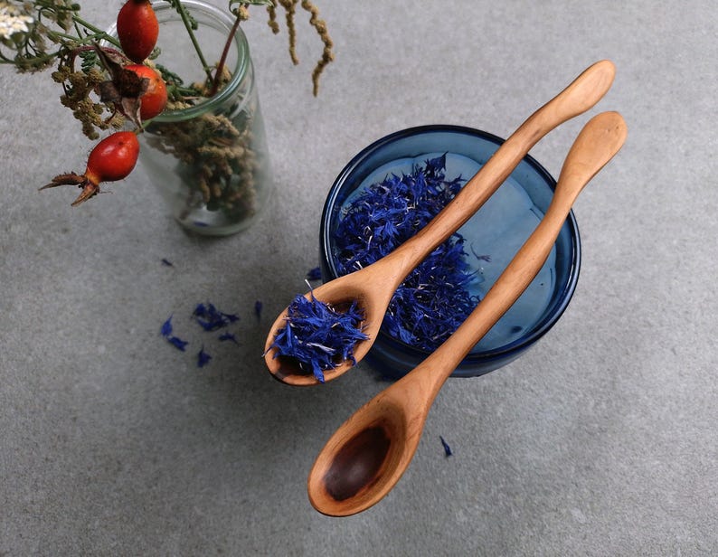 May include: Two light brown wooden spoons with curved handles. One spoon rests inside a dark blue glass bowl filled with dried blue cornflower petals, while the other spoon lies beside it. A clear glass vase with red rose hips is nearby.