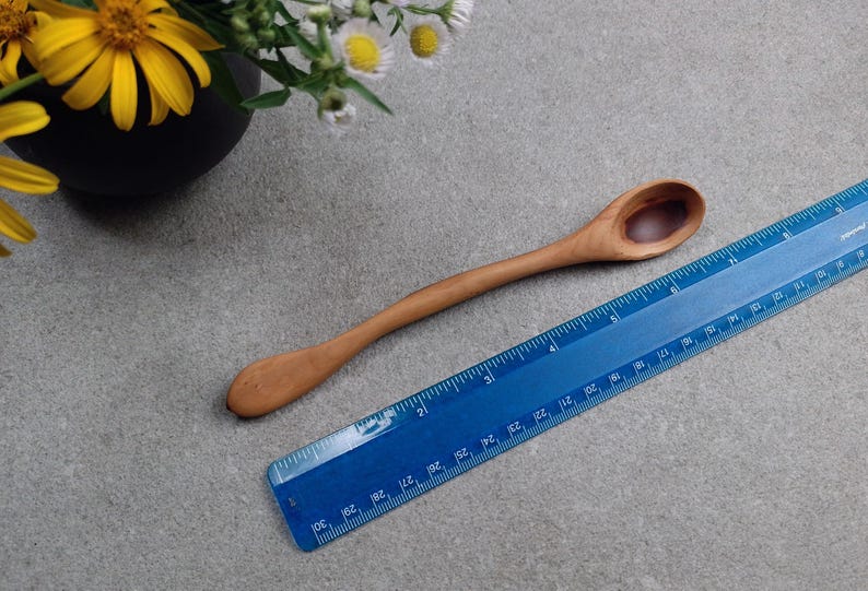 May include: A light brown wooden spoon with a smooth, curved handle and a small, oval-shaped bowl. The spoon is positioned next to a blue plastic ruler that shows the object is approximately 7.5 inches in total length from end to end.