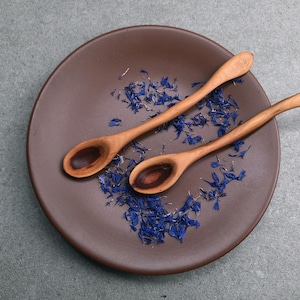 May include: Two light brown wooden spoons with curved handles resting on a dark brown circular plate. The plate is scattered with small, dried blue cornflower petals, creating a contrast between the natural wood grain and the vibrant blue floral elements.