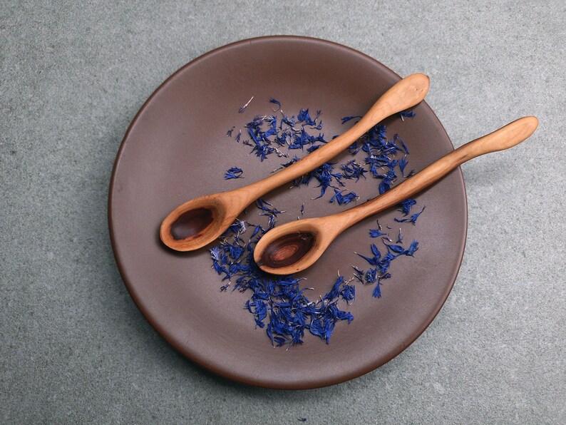 May include: Two light brown wooden spoons with curved handles resting on a dark brown circular plate. The plate is scattered with small, dried blue cornflower petals, creating a contrast between the natural wood grain and the vibrant blue floral elements.