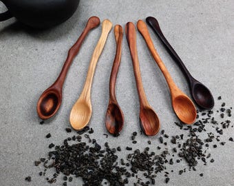 May include: A set of six wooden spoons with long, slender, curved handles, arranged in a fan shape. The spoons feature varying natural wood tones ranging from light tan to deep dark brown, each with a smooth, polished finish and oval bowls.