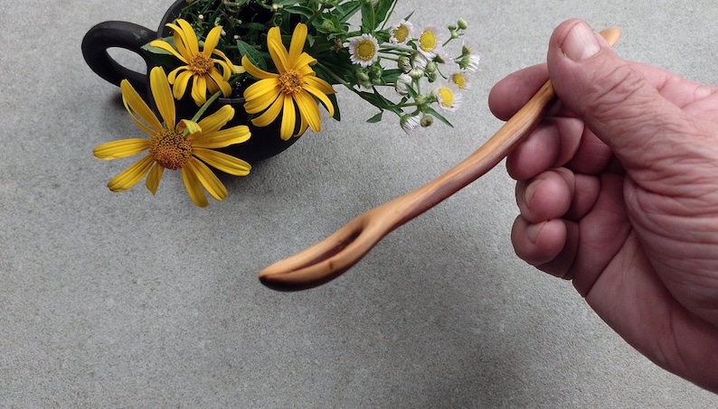 May include: A light-colored wooden spoon with a slender, curved handle and a small, elongated bowl. The wood grain is visible, showing natural variations in tone from light tan to a deeper reddish-brown along the side of the handle.