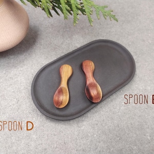 May include: Two small wooden spoons on a black oval dish. The spoons are made of light and dark wood. The text "SPOON E" is printed on the surface below the dish.