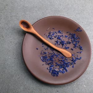 May include: A small, smooth, light-brown wooden spoon resting diagonally across a round, matte, dark-brown ceramic plate. Scattered across the surface of the plate are numerous dried, vibrant blue cornflower petals, creating a contrast.