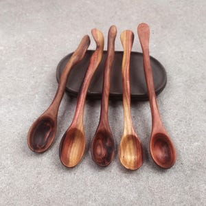 May include: Five long-handled wooden spoons with visible grain patterns in varying shades of reddish-brown. The spoons feature smooth, rounded bowls and slender, slightly curved handles, arranged in a row resting on a dark oval base.