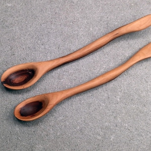 May include: Two long-handled wooden spoons with smooth, curved stems. The bowls of the spoons feature a distinct, dark brown, circular grain pattern that contrasts with the lighter, warm-toned wood of the handles and outer edges.