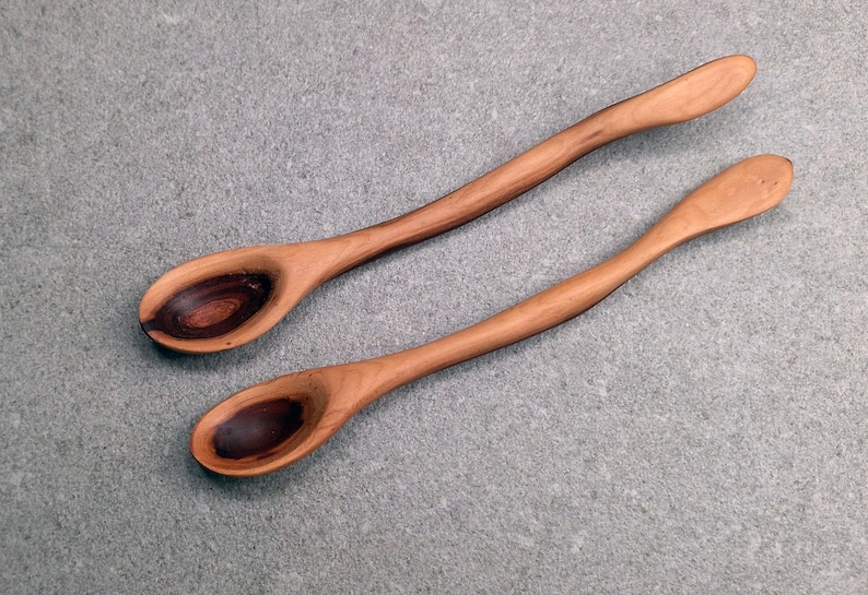 May include: Two long-handled wooden spoons with smooth, curved stems. The bowls of the spoons feature a distinct, dark brown, circular grain pattern that contrasts with the lighter, warm-toned wood of the handles and outer edges.
