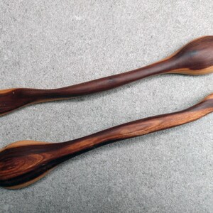 May include: Two handcrafted wooden spoons featuring a distinct two-tone color palette with deep dark brown and lighter tan wood grain patterns. Each spoon has a long, curved handle and a smooth, rounded bowl, showcasing natural wood textures.